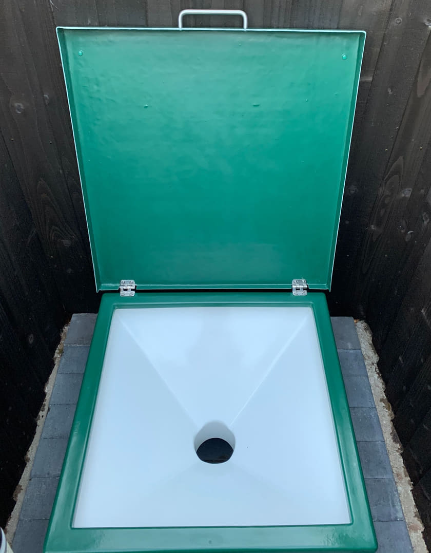 The chemical waste emptying point: a green fibreglass unit with its lid propped open showing a white moulded funnel inside, set on grey block paving within a dark stained timber screen.
