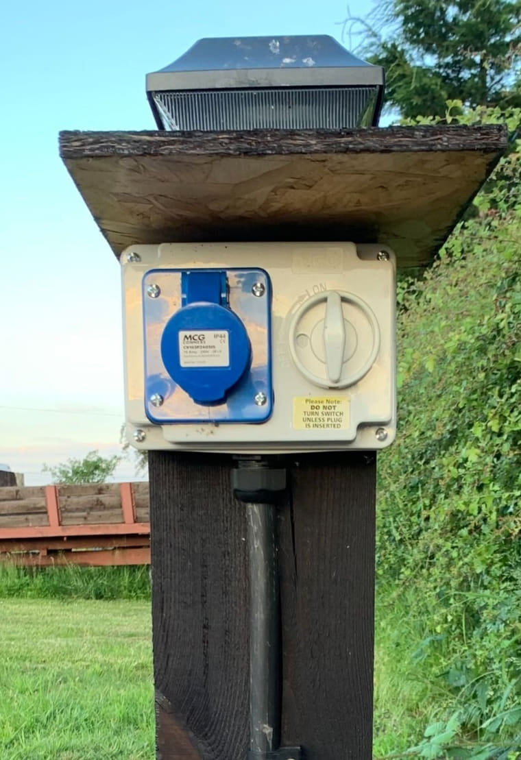 A cream weatherproof enclosure holding a blue 16 amp commando socket and a rotary isolator, mounted on a black stained timber post under a small timber rain hood with a solar light on top.