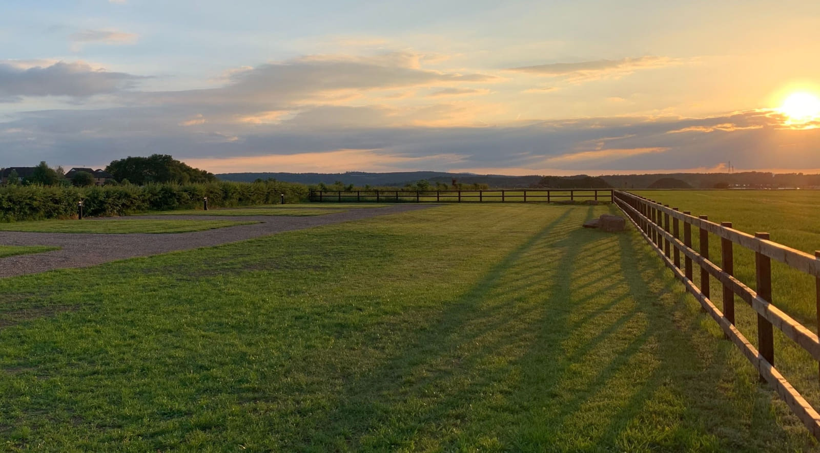 Low evening sun flaring behind a new timber post and rail fence, with the mown grass pitch area, a gravel track and solar bollard lights along the hedge.