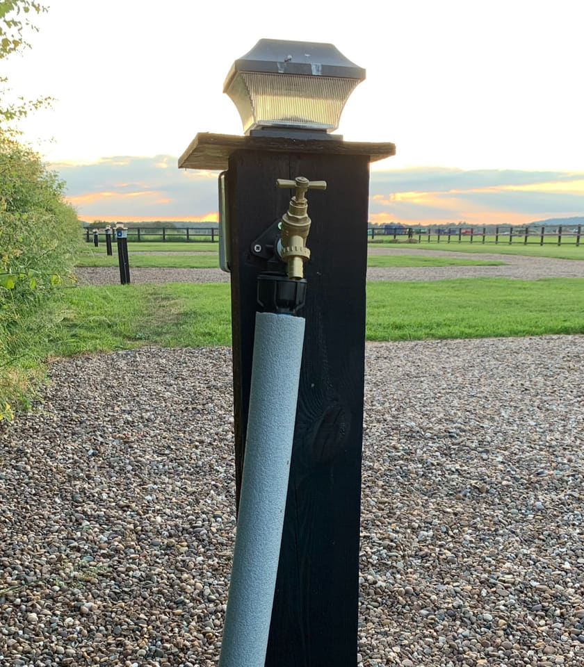 A brass drinking water tap on a black stained timber post with a solar light on its cap and the supply pipe lagged, standing on gravel beside the pitching area at sunset.